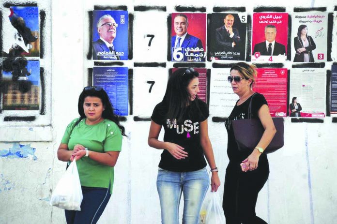 Women walk past candidates' posters and designated numbers a day before the start of presidential election in Tunis, Tunisia, Saturday, Sept. 14, 2019. AP