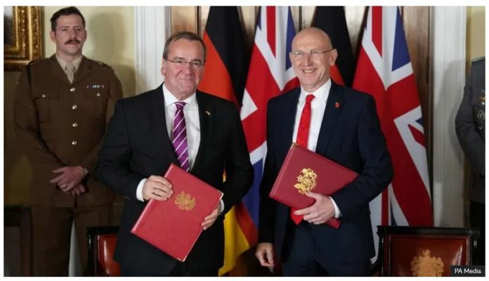 UK United Kingdom’s Defence Secretary John Healey and Germany’s Defence Minister Boris Pistorius smile, standing in front of the Union Jack and German flags, holding red binders. BBC