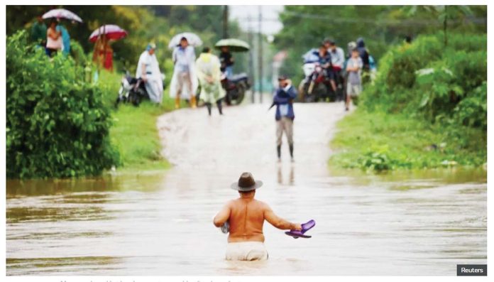 A shirtless man wearing a brown hat and holding his flip flops in one hand wades across waist-deep water while people watch from the other side of an inundated road in El Progreso, Honduras. Many roads and bridges became impassable after days of rain. REUTERS