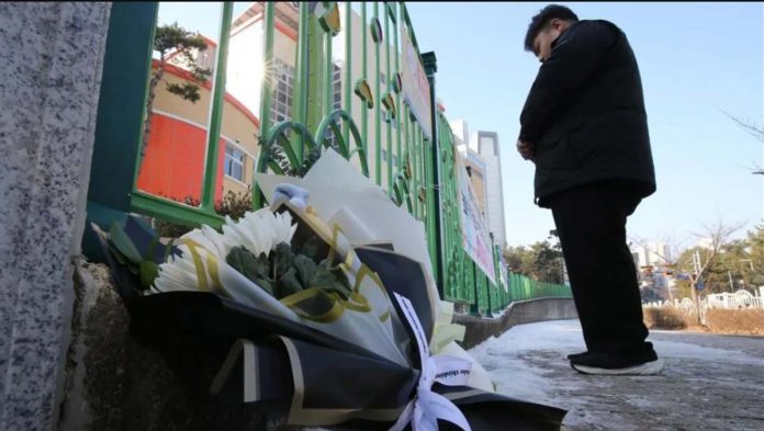 South-Korea A man stands with his hands crossed in front of a school gate. Bouquets of flowers lay on the ground beside him. Locals laid flowers at the school’s gate to mourn an eight-year-old student. NEWS1