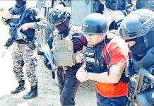 Ecuadorean drug lord “Fito” wears a hard hat and an orange t-shirt underneath a bullet-proof vest. He is escorted by several people in black and camouflage clothes, also wearing bullet-proof vests. SNAI