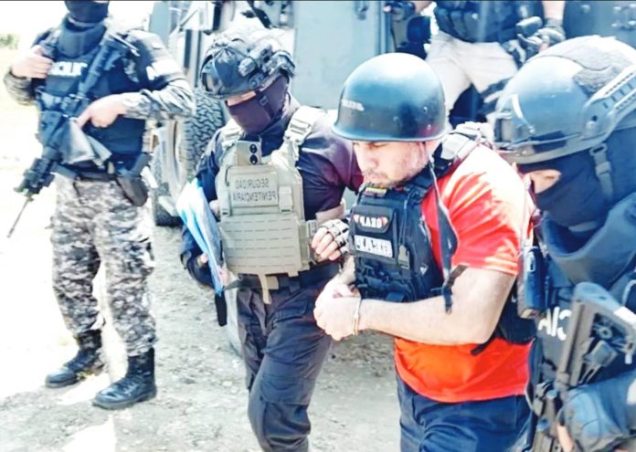 Ecuadorean drug lord “Fito” wears a hard hat and an orange t-shirt underneath a bullet-proof vest. He is escorted by several people in black and camouflage clothes, also wearing bullet-proof vests. SNAI