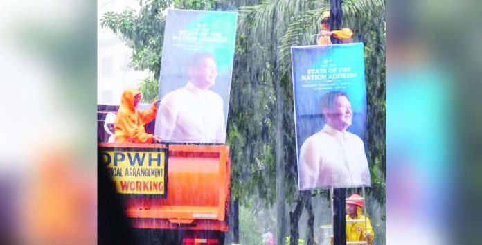 PBBM copy_superimposed Undeterred by the heavy downpour, Department of Public Works and Highways personnel pressed on with installing tarpaulins promoting President Ferdinand “Bongbong” Marcos Jr.’s upcoming State of the Nation Address. They work full-force despite weather disruptions brought by the habagat. PHOTO COURTESY OF DWAR 1494