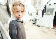 A Palestinian child, wet from rain, looks into the camera, surrounded by soaked tents and wet and muddy ground. BBC