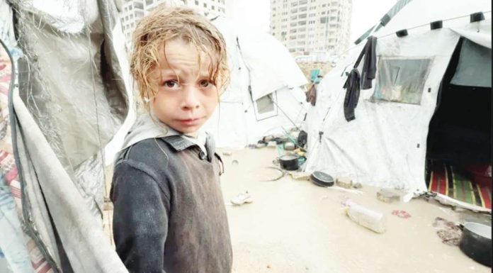 Gaza A Palestinian child, wet from rain, looks into the camera, surrounded by soaked tents and wet and muddy ground. BBC