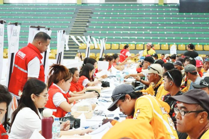 estember Regional Director Joselito G. Estember (standing, left) of the Department of Social Welfare and Development Region 6 oversees the distribution of cash relief to tricycle driver beneficiaries at the Passi City Arena in Passi City, Iloilo on Wednesday, April 8, 2026. He personally monitors the payout flow and implements an action plan to expedite the process for 2,600 transport sector workers affected by the ongoing economic crisis. DSWD REGION 6 PHOTO