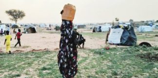 A woman, with her back to the camera and carrying a drum on her head, is walking in a camp, with children seen not far from her. The conflict in Sudan has forced millions of people to flee their homes. REUTERS