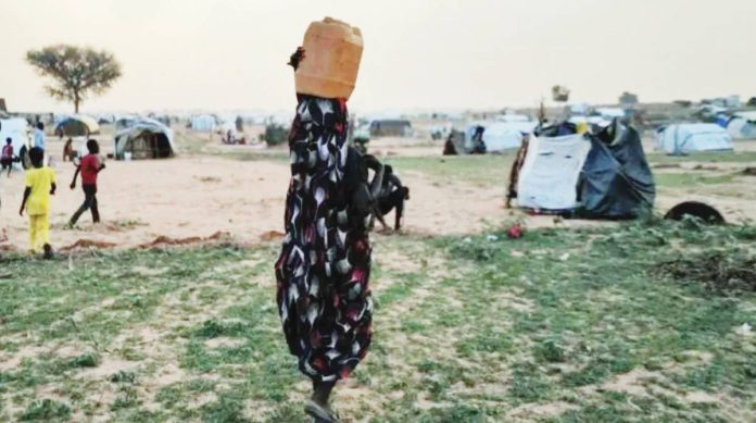 Sudan A woman, with her back to the camera and carrying a drum on her head, is walking in a camp, with children seen not far from her. The conflict in Sudan has forced millions of people to flee their homes. REUTERS