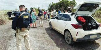A federal highway policeman in Brazil stands a few yards from a white dusty car that has its boot open. In the background, an officer can be seen talking to a woman, while a row of about a dozen people is lined up on the roadside. INTERPOL