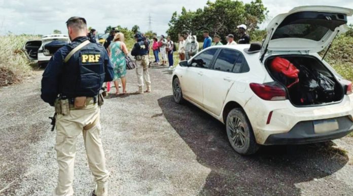 Global A federal highway policeman in Brazil stands a few yards from a white dusty car that has its boot open. In the background, an officer can be seen talking to a woman, while a row of about a dozen people is lined up on the roadside. INTERPOL
