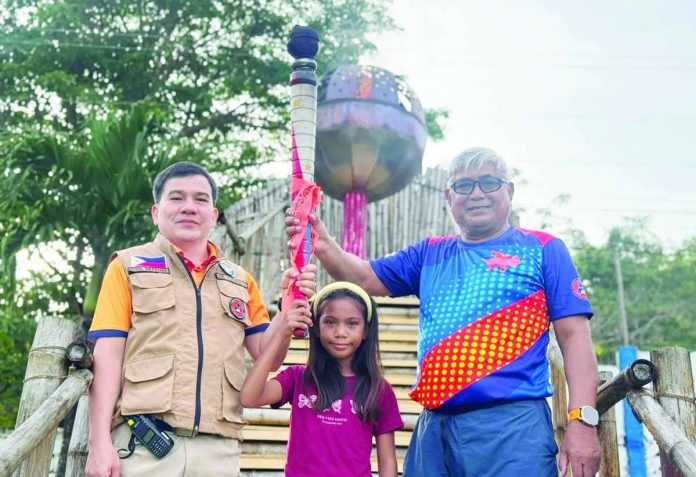 Office of Civil Defense Regional Office 6 Director Raul E. Fernandez (left) and Broderick G. Train, head of the Provincial Disaster Risk Reduction and Management Office of Antique, pose with 12-year-old Cherry Ann Mateo after she received a Plaque of Heroism and cash assistance during the opening of Rescuelympics 2026 on July 28. Cherry Ann was recognized for her courageous act of swimming for nearly five hours to seek help after a motorboat capsized off Batbatan Island, leading to the successful rescue of all its passengers. PDRRMO ANTIQUE PHOTO