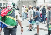 A masked man draped in a South African flag stands in the street as onlookers gather behind him, holding a slingshot in one hand and a stick in the other. Anti-migrant protesters in South Africa demand that all undocumented foreign nationals leave the country. GETTY IMAGES