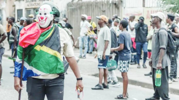 A masked man draped in a South African flag stands in the street as onlookers gather behind him, holding a slingshot in one hand and a stick in the other. Anti-migrant protesters in South Africa demand that all undocumented foreign nationals leave the country. GETTY IMAGES