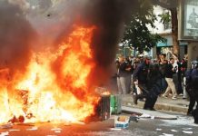 A large fire burns on a road in Paris beside which five officers in riot gear walk past a crowd observing the flames. Hundreds of thousands of protesters take to the streets across France. REUTERS