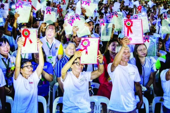 neljoy_land_titles_copy_compressed_resized Agrarian reform beneficiaries raise their newly awarded land titles during the Department of Agrarian Reform (DAR) – Region 6’s mass distribution ceremony at the Iloilo National High School in La Paz, Iloilo City. DAR distributed 11,965 e-titles covering 18,598 hectares to 10,998 farmers across Panay and Guimaras islands, strengthening individual land ownership and tenure security among beneficiaries. ALLEN ANIMAS/PN